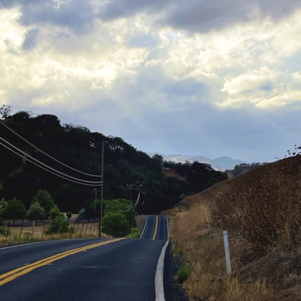 Photo of an empty country road surrounded by hills and trees, with a dramatic cloudy sky in the background