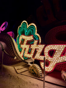 Photo of part of a vintage neon sign for the Fitzgerald casino, with a green shamrock next to lettering in script type, edged in red and filled with round white bulbs. It is resting on the ground next to a semi-buried gold coin marked with the letter F.